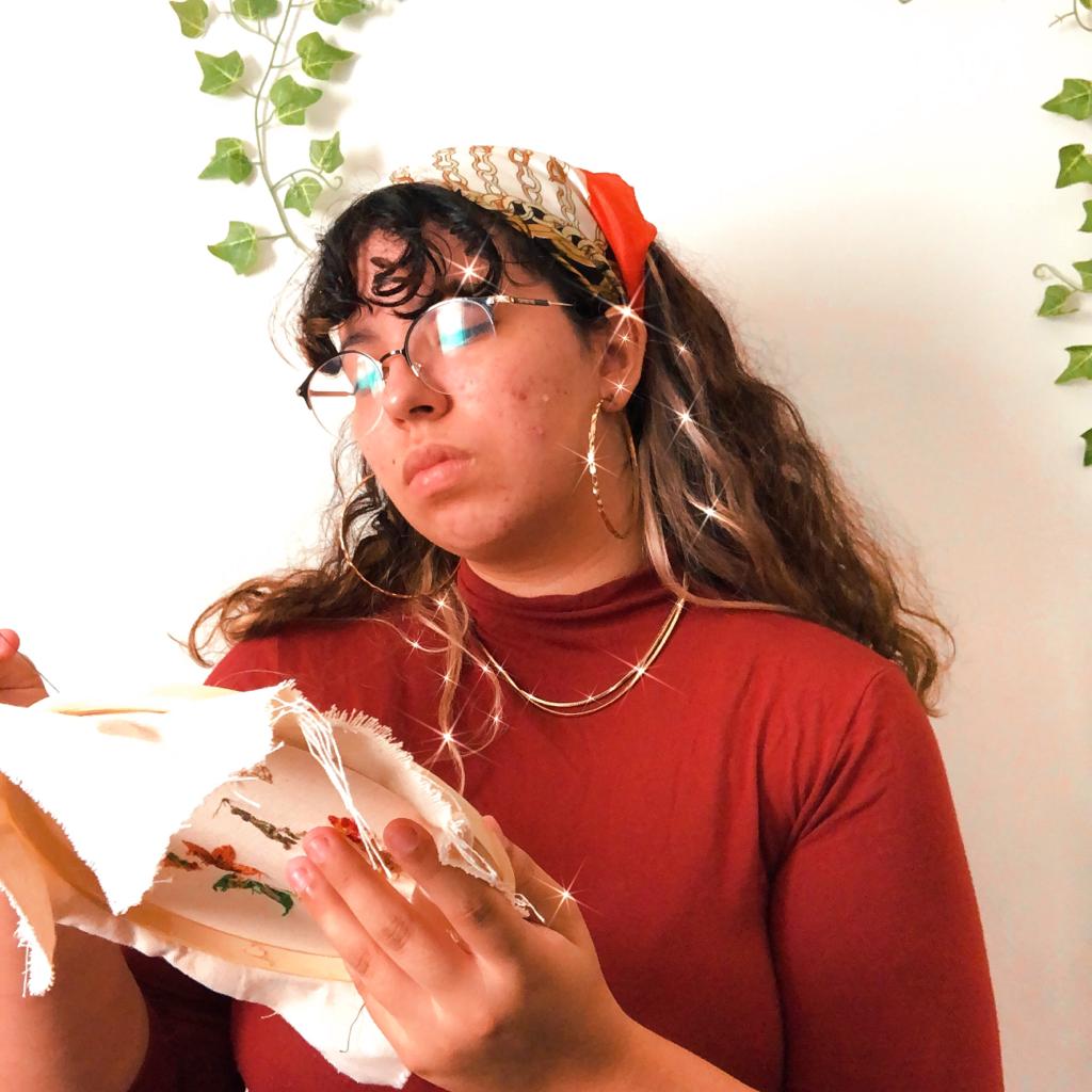 Raíssa, dona da loja, em um cenario com plantas, bordando uma das peças feitas á mão